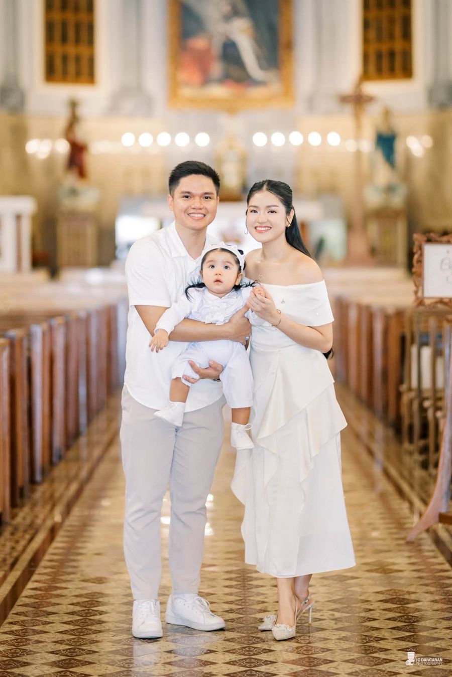 Parents holding their baby in a church aisle, all dressed in white